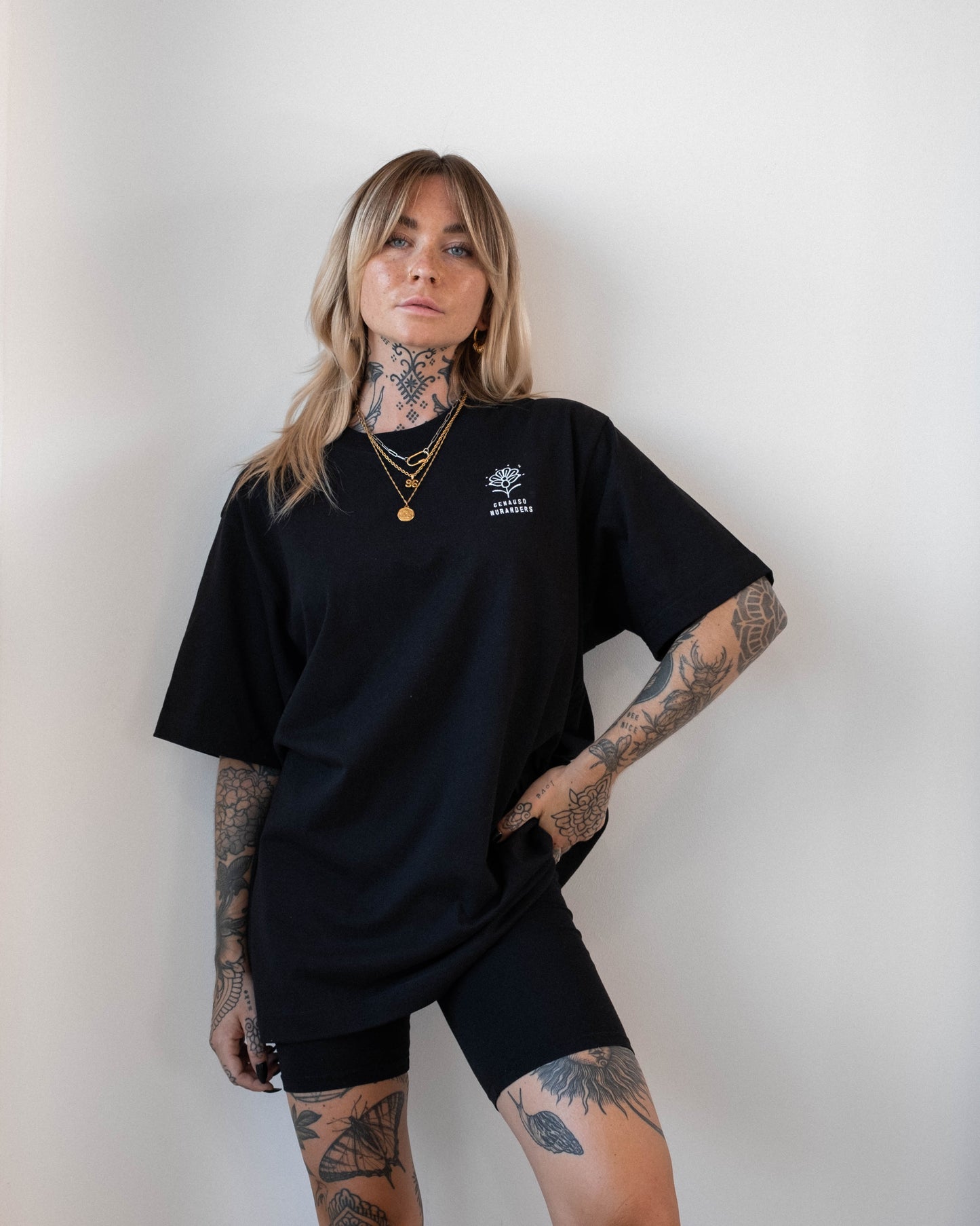 Schwarzes T-Shirt mit weißem Frontprint, bestehend aus einer grafischen Blume und dem Text „GENAUSO NUR ANDERS“. Die tätowierte Person steht seitlich zur Kamera und trägt das Shirt locker über einer schwarzen Shorts.