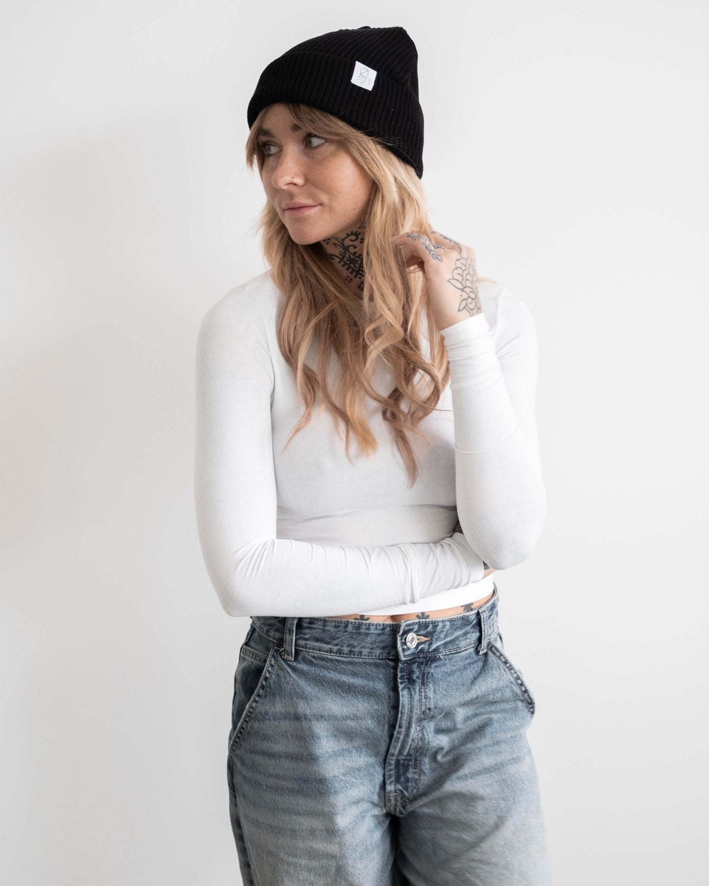 Frau mit schwarzer Beanie und weißem Langarmshirt steht mit verschränkten Armen vor hellem Hintergrund. Auf der Beanie ist ein kleines weißes Logoetikett sichtbar. Ihr blondes Haar fällt locker über die Schultern. Sie blickt zur Seite.