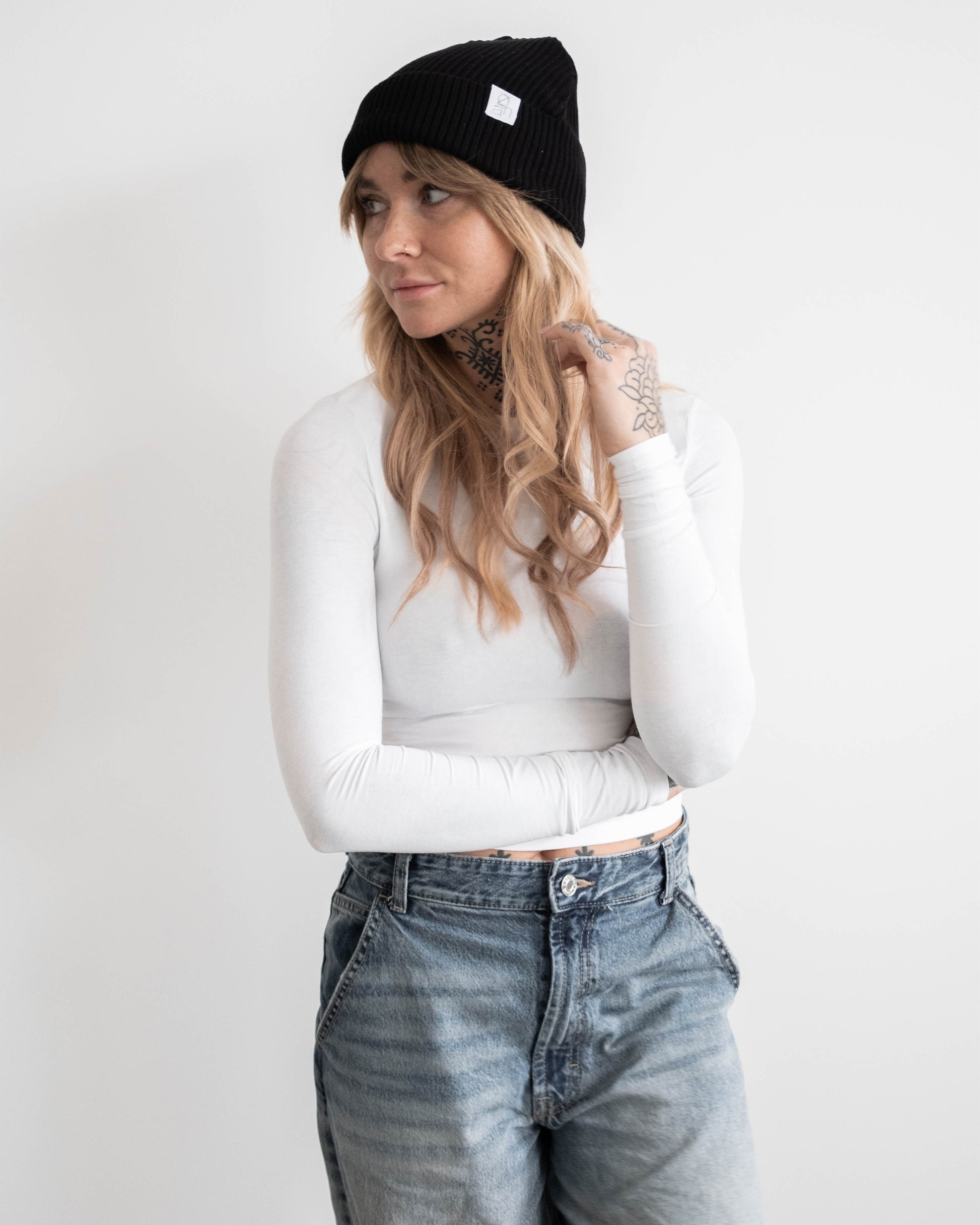Frau mit schwarzer Beanie und weißem Langarmshirt steht mit verschränkten Armen vor hellem Hintergrund. Auf der Beanie ist ein kleines weißes Logoetikett sichtbar. Ihr blondes Haar fällt locker über die Schultern. Sie blickt zur Seite.