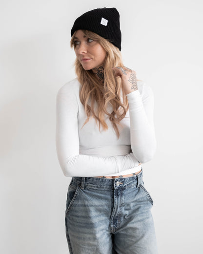 Frau mit schwarzer Beanie und weißem Langarmshirt steht mit verschränkten Armen vor hellem Hintergrund. Auf der Beanie ist ein kleines weißes Logoetikett sichtbar. Ihr blondes Haar fällt locker über die Schultern. Sie blickt zur Seite.