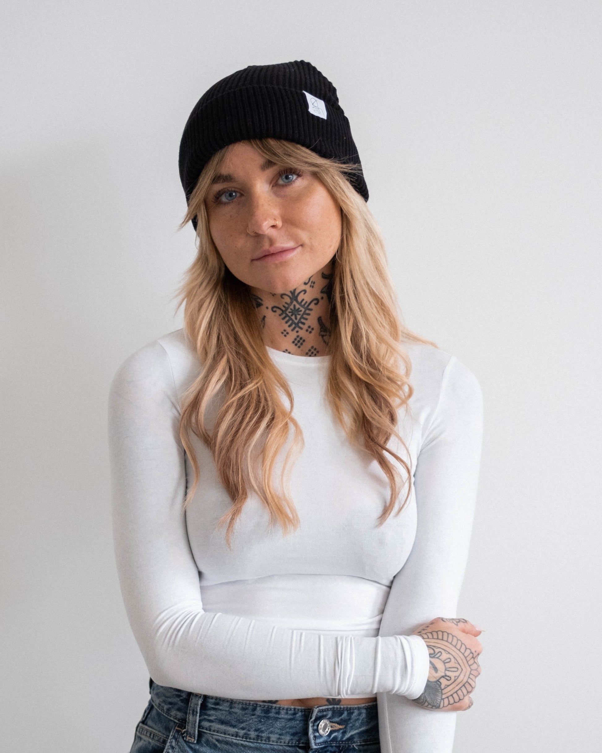 Nahaufnahme einer Frau mit schwarzer Beanie-Mütze mit weißem Logoetikett. Sie trägt ein weißes, eng anliegendes Langarmshirt und eine Jeans. Ihre Arme sind verschränkt, sie schaut direkt in die Kamera. Der Hintergrund ist hell und neutral.