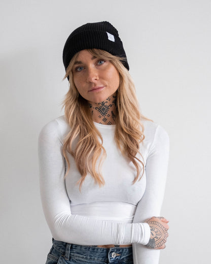 Nahaufnahme einer Frau mit schwarzer Beanie-Mütze mit weißem Logoetikett. Sie trägt ein weißes, eng anliegendes Langarmshirt und eine Jeans. Ihre Arme sind verschränkt, sie schaut direkt in die Kamera. Der Hintergrund ist hell und neutral.
